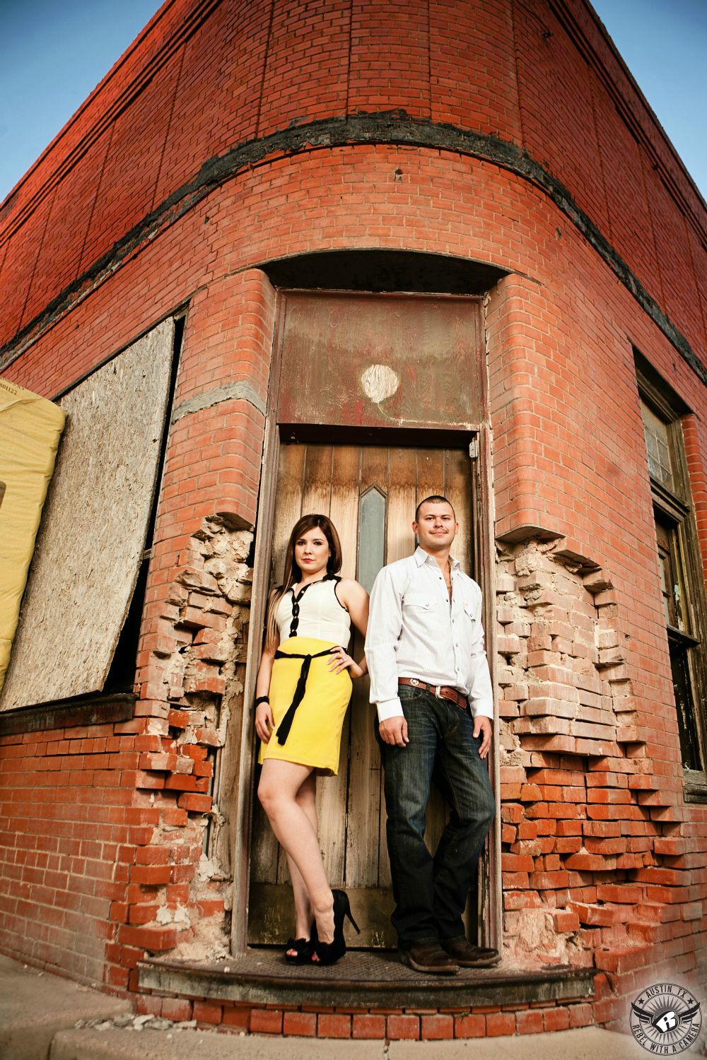 hispanic engagement couple in white tank top blouse with a knee length yellow skirt and white cowboy shirt with dark blue jeans standing in door way of a decrepit red brick building in granger texas 